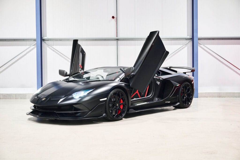 A black sports car with scissor doors raised is parked indoors on a light-colored floor. The car, available for hire, features red brake calipers and a rear spoiler.