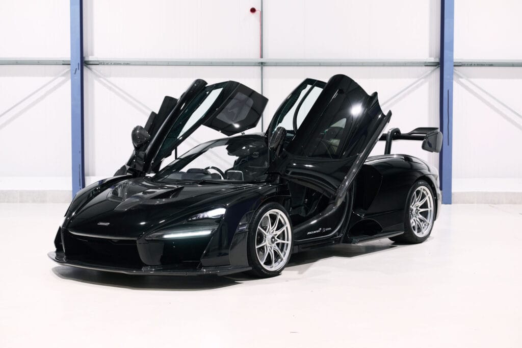 A black mclaren sports car with both doors open, parked inside a showroom with a white floor and walls.