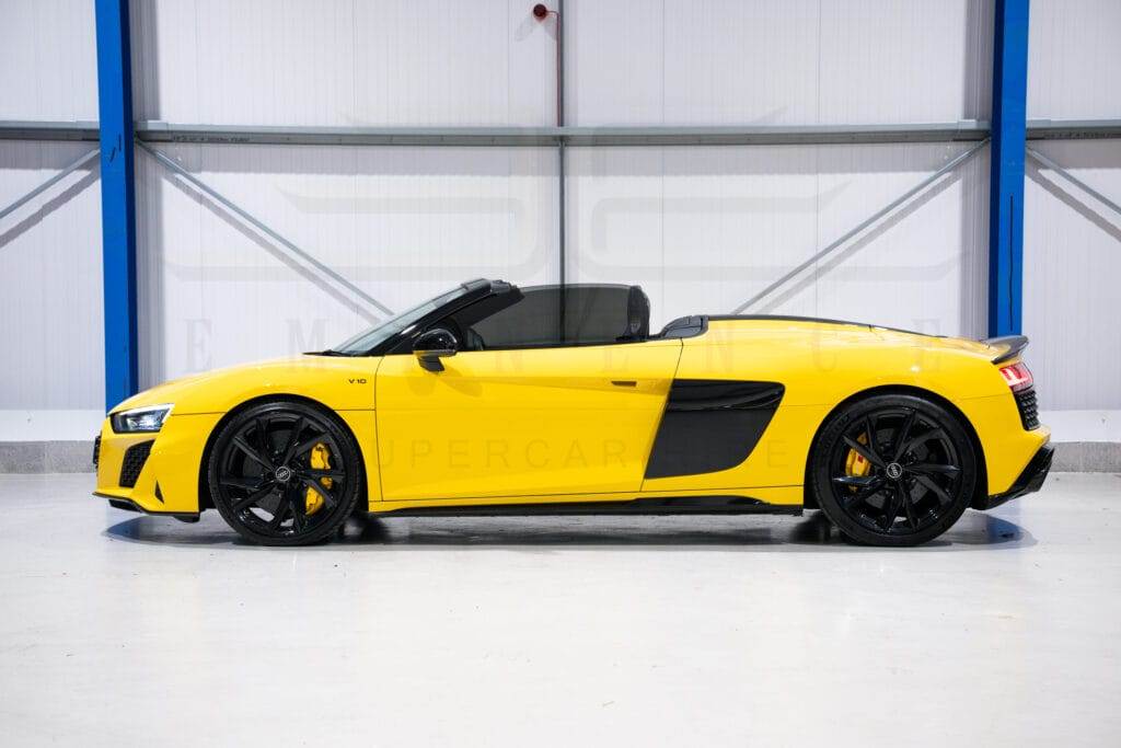 Side view of a yellow convertible sports car with black accents and black wheels parked in a garage, available for hire.