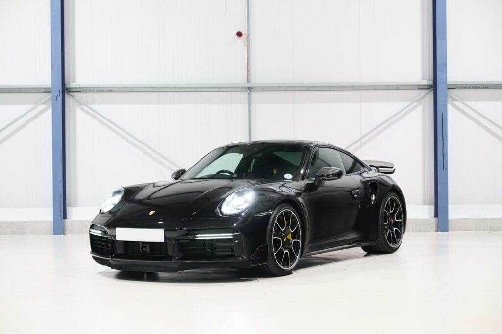 A sleek black sports car is parked in a large, well-lit indoor space with a white wall and blue support beams in the background, waiting for someone to hire it for their next adventure.