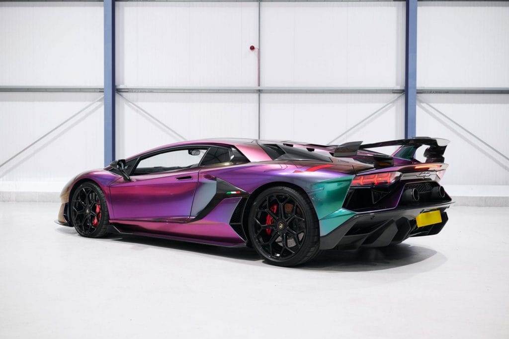 A multicolored, metallic car with a sleek design is parked indoors against a plain backdrop. Available for hire, the car features a large rear spoiler and black rims.