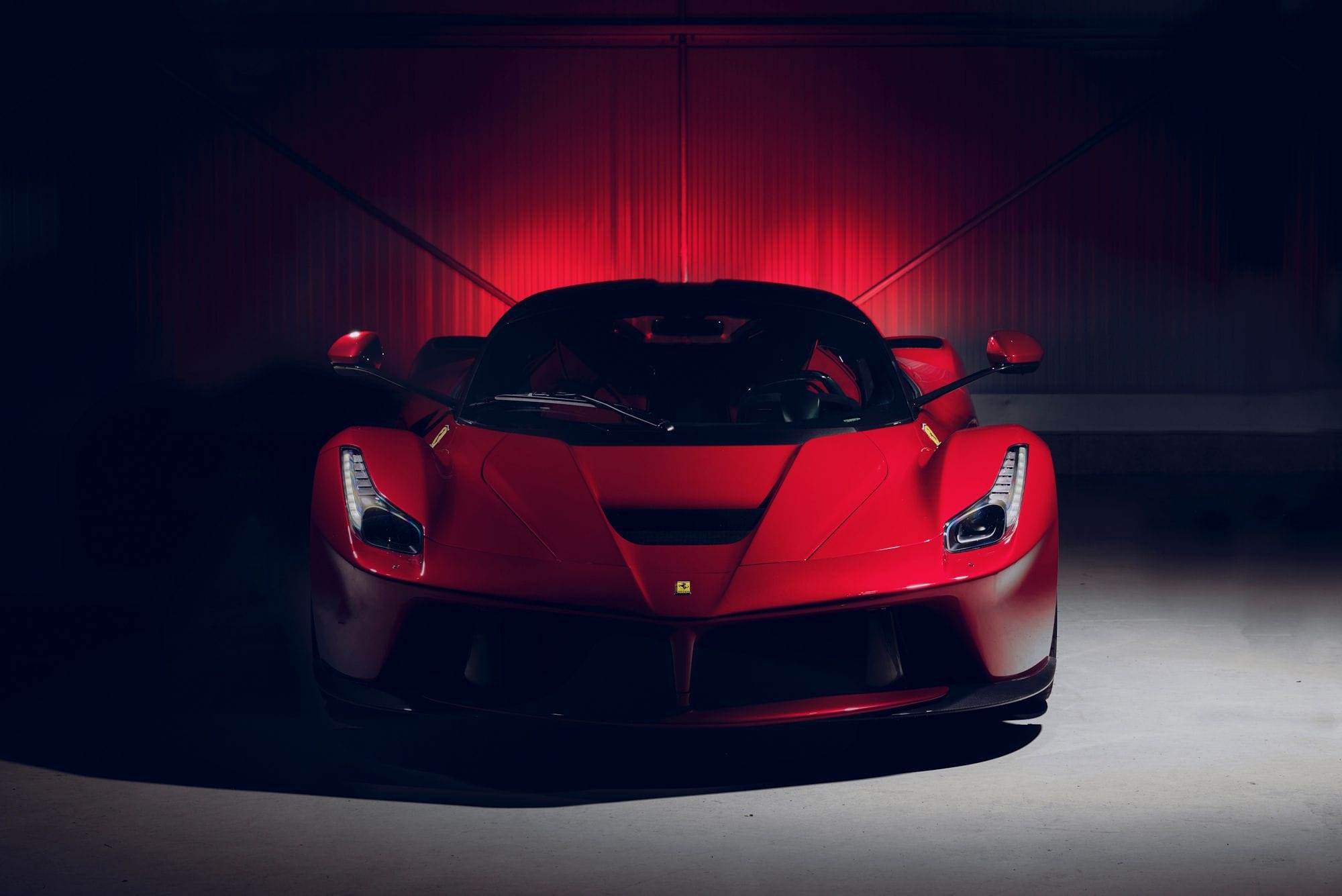 Front view of a red sports car with illuminated headlights, set against a dark background with a red light halo effect behind the vehicle, ready for hire.